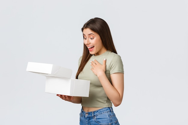Koncepcja dostawy, stylu życia i żywności. Zaskoczona szczęśliwa kobieta otrzymuje pudełko-niespodziankę z deserami, pysznymi słodkimi ciastami i babeczkami, zdumiona w środku, stojąca na białym tle.