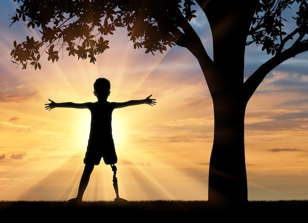 Koncepcja dzieci niepełnosprawnych. Szczęśliwy niepełnosprawny chłopiec z protezą nogi stojący w pobliżu drzewa o zachodzie słońca
