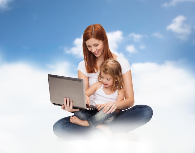 Zdjęcie koncepcja dzieciństwa, rodzicielstwa, rodziny, ludzi i technologii - szczęśliwa matka z uroczą dziewczynką z laptopem na tle błękitnego nieba i chmur