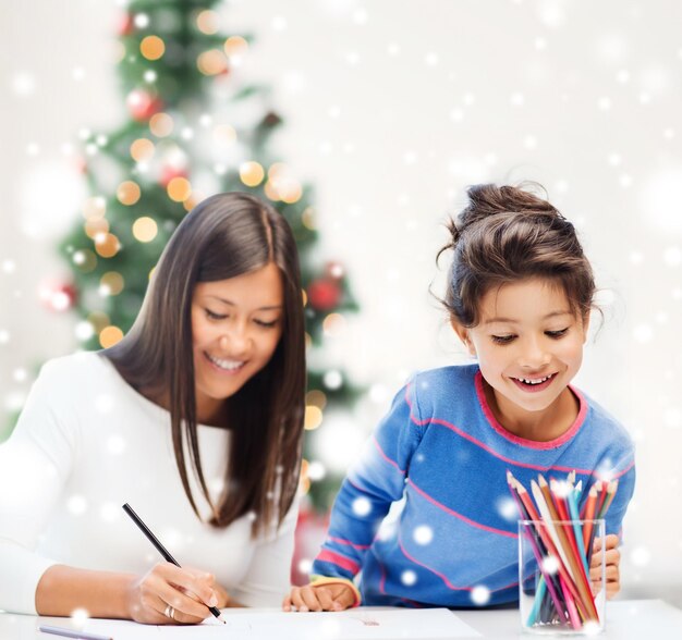 koncepcja dzieciństwa, rodziny, świąt Bożego Narodzenia i ludzi - uśmiechnięta dziewczynka i matka lub nauczyciel rysujący kredkami w pomieszczeniu
