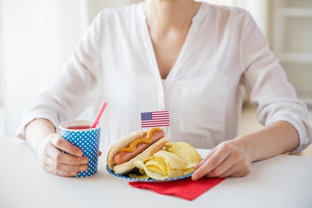 Zdjęcie koncepcja dzień niepodległości, świętowanie, patriotyzm i święta - zbliżenie kobiety jedzącej hot doga z dekoracją flagi amerykańskiej i chipsami ziemniaczanymi, pijące sok i świętujące 4 lipca na imprezie domowej