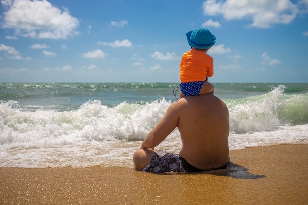 koncepcja dzień ojca. tata z dzieckiem na ramionach siedzą na piasku i patrzą na morze.