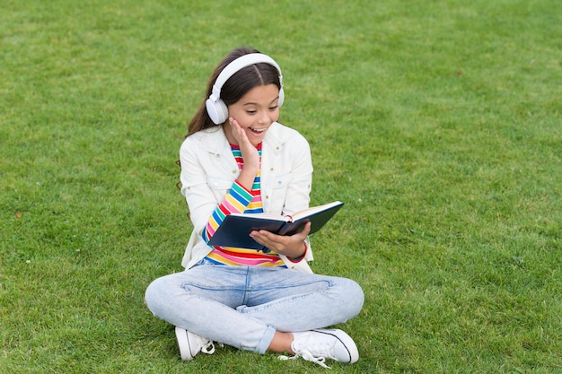 Koncepcja edukacji i czytania rozwój wyobraźni ładna dziewczyna czyta książkę szczęśliwa dziewczynka w wieku przedszkolnym z książką na szkolnym podwórku powrót do szkoły pracowite dziecko w słuchawkach