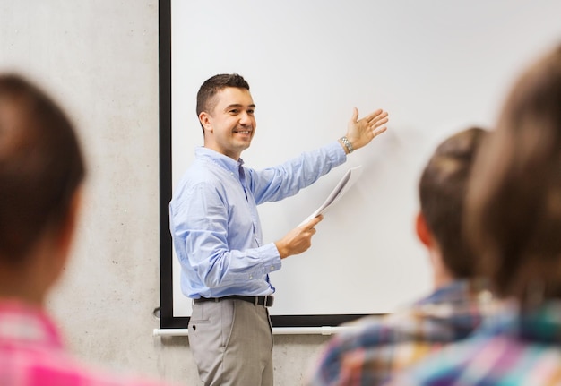 koncepcja edukacji, liceum, technologii i ludzi - uśmiechnięty nauczyciel z notatnikiem, laptop stojący przed uczniami i pokazujący coś na białej tablicy w klasie
