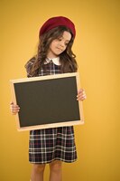 Koncepcja edukacji szczęśliwa dziewczyna we francuskim berecie tablica reklamowa do promocji zakupy szkolne sprzedaż dziecko na żółtym tle z powrotem do szkoły mała dziewczynka z miejscem na kopię tablicy szkolnej