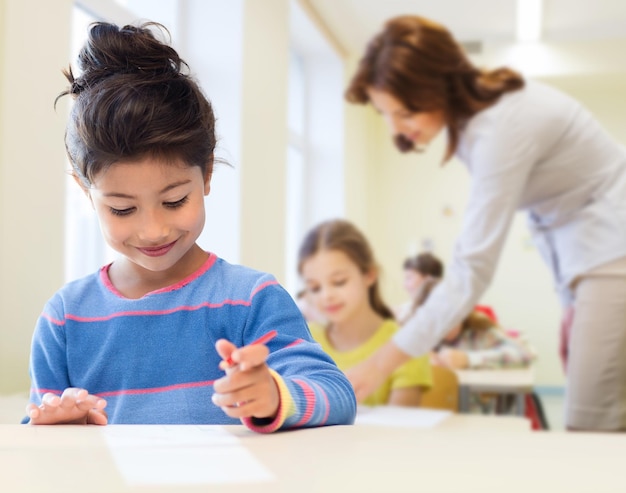 koncepcja edukacji, szkoły podstawowej i dzieci - szczęśliwa mała studentka z piórem i papierem do pisania na tle klasy i nauczyciela