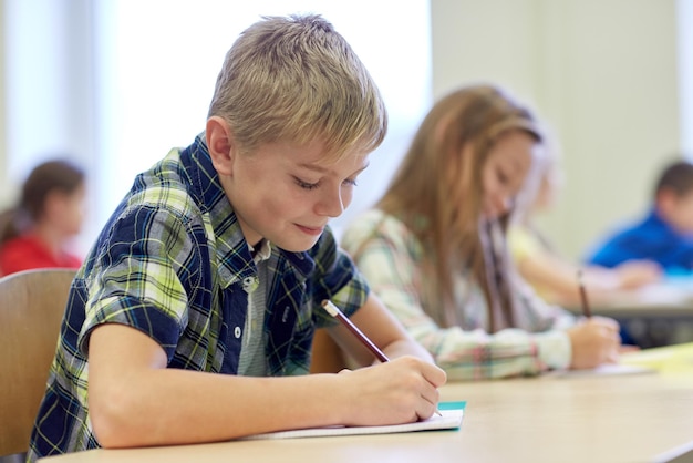 koncepcja edukacji, szkoły podstawowej, uczenia się i ludzi - grupa dzieci w wieku szkolnym z długopisami i zeszytami pisząca test w klasie