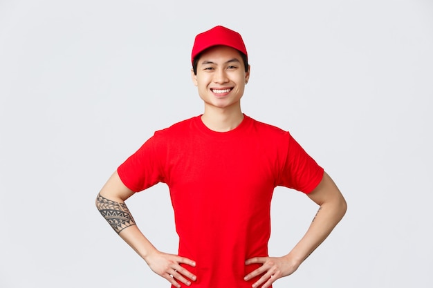 Koncepcja ekspresowej dostawy, wysyłki i logistyki. Wesoły pewny siebie azjatycki kurier w czerwonej czapce i koszulce, stojący entuzjastycznie z rękami na biodrach, uśmiechnięty, dostarczający zamówienie klientowi na czas.