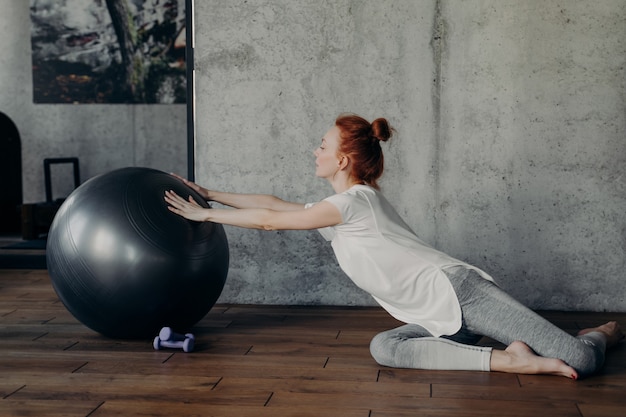 Koncepcja fitness i gimnastyki. Młoda wesoła wysportowana kobieta z rudymi włosami w kok w odzieży sportowej robi ćwiczenia równowagi z fitball na drewnianej podłodze obok betonowej ściany w siłowni, ćwiczenia w pomieszczeniu