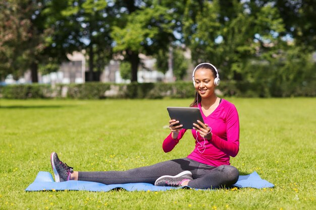 koncepcja fitness, parku, technologii i sportu - uśmiechnięta afroamerykanka z komputerem typu tablet i słuchawkami na macie na zewnątrz