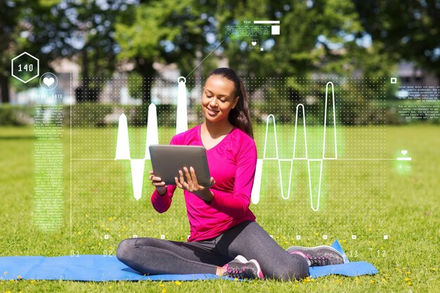 koncepcja fitness, parku, technologii i sportu - uśmiechnięta Afroamerykanka z komputerem typu tablet PC siedzi na macie na zewnątrz