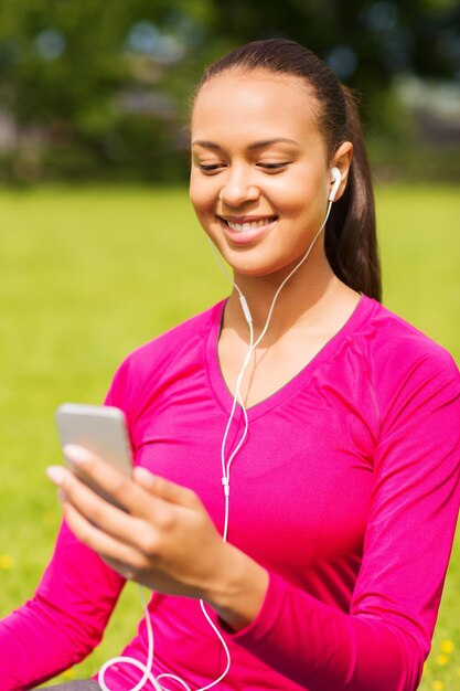 koncepcja fitness, parku, technologii i sportu - uśmiechnięta Afroamerykanka ze smartfonem i słuchawkami siedzi na macie na zewnątrz