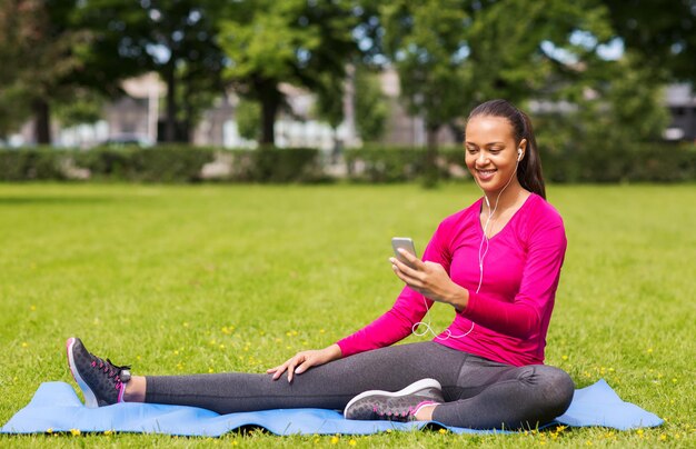 koncepcja fitness, parku, technologii i sportu - uśmiechnięta Afroamerykanka ze smartfonem i słuchawkami siedzi na macie na zewnątrz