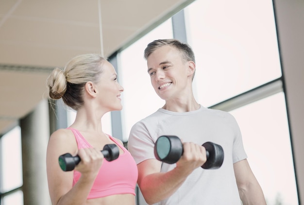 koncepcja fitness, sportu, ćwiczeń i diety - uśmiechnięta młoda kobieta i osobisty trener z hantlami w siłowni