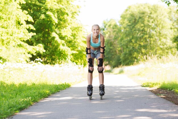 koncepcja fitness, sportu, lata, jazdy na rolkach i zdrowego stylu życia - szczęśliwa młoda kobieta w rolkach i sprzęcie ochronnym jeżdżąca na świeżym powietrzu