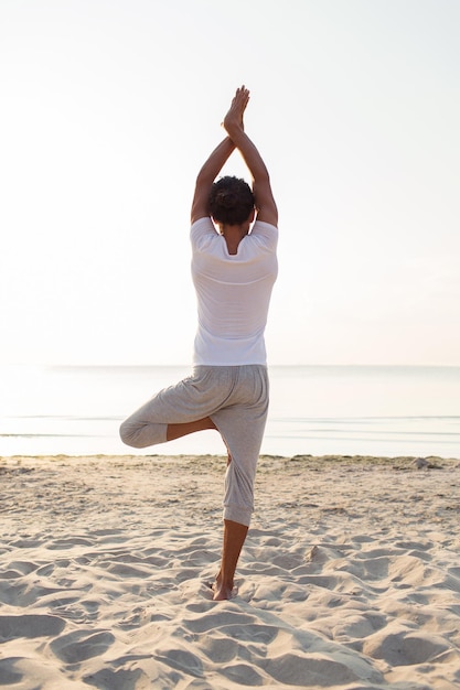 koncepcja fitness, sportu, ludzi i stylu życia - człowiek wykonujący ćwiczenia jogi na piasku na zewnątrz z tyłu