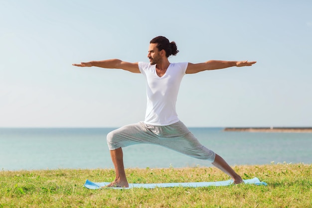 koncepcja fitness, sportu, ludzi i stylu życia - uśmiechnięty mężczyzna wykonujący ćwiczenia jogi na macie na zewnątrz