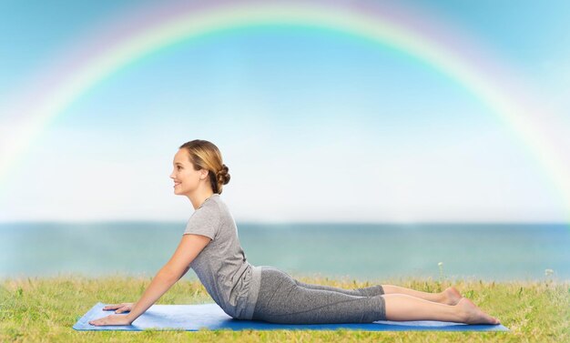 koncepcja fitness, sportu, ludzi i zdrowego stylu życia - kobieta robi jogę w pozie psa na macie na tle błękitnego nieba, tęczy i morza