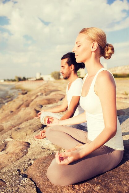 koncepcja fitness, sportu, przyjaźni i stylu życia - uśmiechnięta para wykonująca ćwiczenia jogi siedząca na zewnątrz