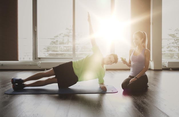 koncepcja fitness, sportu, technologii i ludzi - mężczyzna i kobieta ze smartfonem robią ćwiczenia z deski bocznej na macie w siłowni