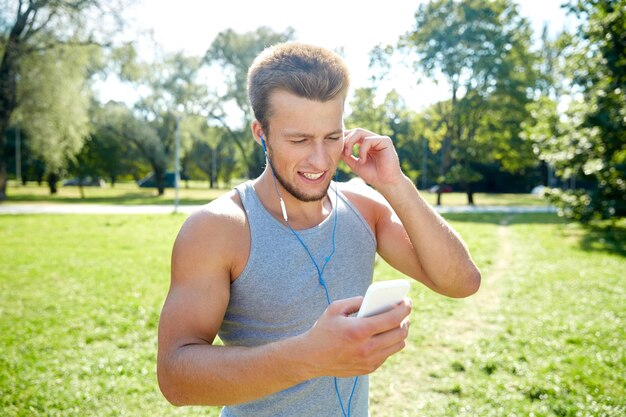 koncepcja fitness, sportu, technologii i stylu życia - szczęśliwy młody człowiek ze smartfonem i słuchawkami do słuchania muzyki w letnim parku