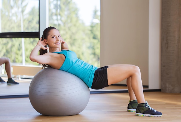 koncepcja fitness, sportu, treningu i ludzi - uśmiechnięta kobieta napina mięśnie brzucha z piłką do ćwiczeń w siłowni