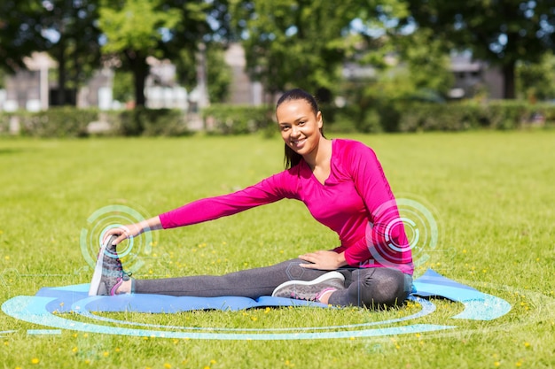 koncepcja fitness, sportu, treningu, parku i stylu życia - uśmiechnięta kobieta rozciągająca nogę na macie na zewnątrz