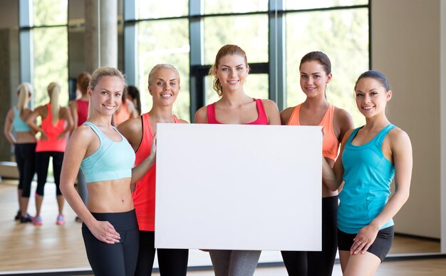 koncepcja fitness, sportu, treningu, siłowni i stylu życia - grupa kobiet z białym pustym billboardem w siłowni