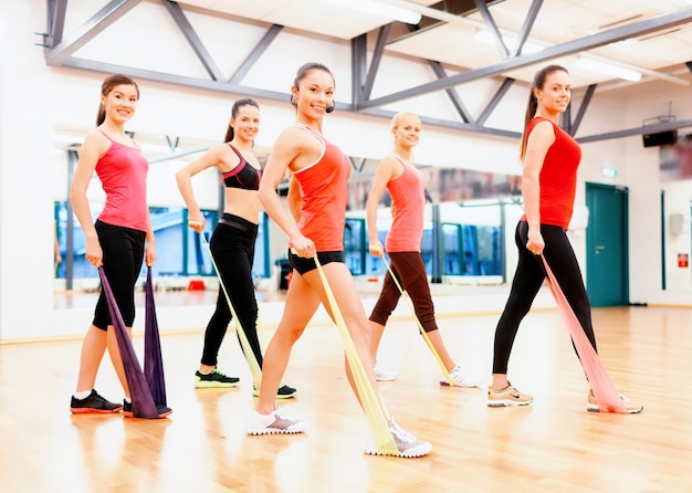 koncepcja fitness, sportu, treningu, siłowni i stylu życia - grupa uśmiechniętych osób z instruktorem ćwiczącym z gumkami na siłowni