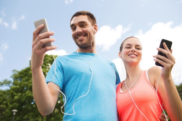 koncepcja fitness, sportu, treningu, technologii i stylu życia - dwie uśmiechnięte osoby ze smartfonami i słuchawkami na zewnątrz