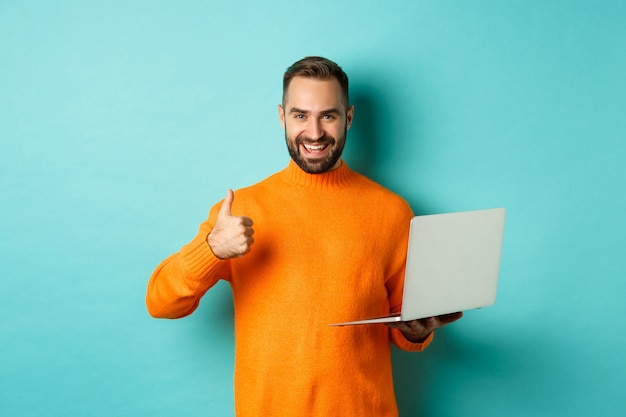 Koncepcja freelance i technologii. Szczęśliwy człowiek w pomarańczowym swetrze, pokazujący kciuk do góry podczas pracy z laptopem, stojący zadowolony na jasnoniebieskim tle.