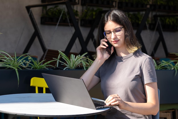 Koncepcja Freelancera Piękna Brunetka Studentka W Okularach Rozmawia Przez Telefon, Używając Laptopa W Kawiarni