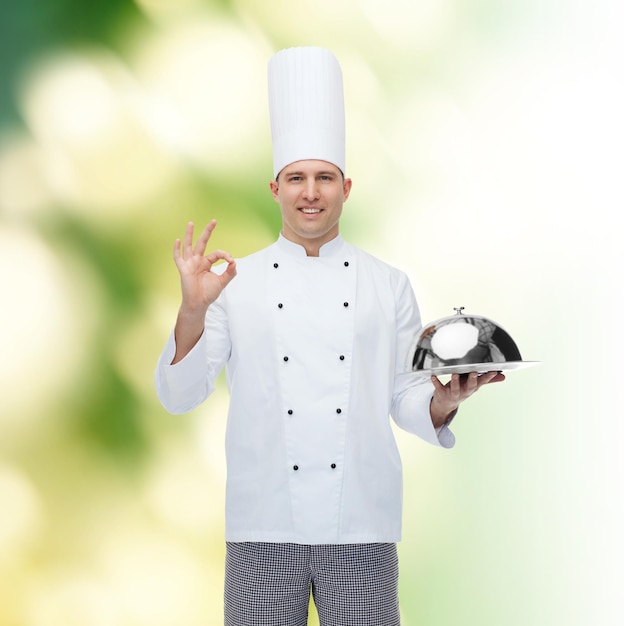 koncepcja gotowania, zawodu, gestów i ludzi - szczęśliwy mężczyzna kucharz kucharz trzyma klosz i pokazuje znak ok na zielonym tle