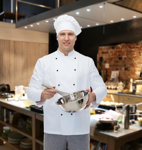 koncepcja gotowania, zawodu i ludzi - szczęśliwy mężczyzna kucharz kucharz trzyma miskę i ubija coś trzepaczką nad kuchnią restauracji