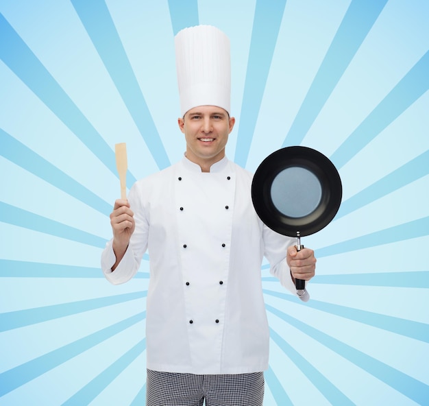koncepcja gotowania, zawodu i ludzi - szczęśliwy mężczyzna kucharz kucharz trzyma patelnię i łopatkę na niebieskim tle promieni wybuchu