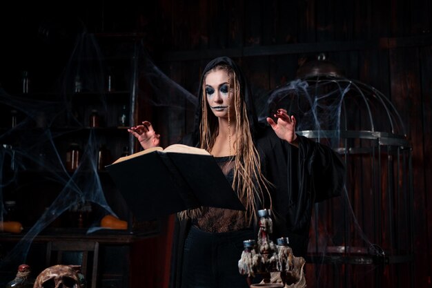 Koncepcja Halloween. Czarownica ubrana w czarny kaptur stojąca w ciemnym lochu używa magicznej księgi do wyczarowania magicznego zaklęcia. Kobieca czarodziejka nekromanta gotyckie wnętrze z klatką, pajęczyna