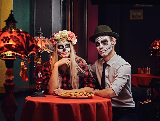 Koncepcja Halloween i Muertos. Młoda atrakcyjna para z nieumarłym makijażem jedząca nachos podczas randki w meksykańskiej restauracji.