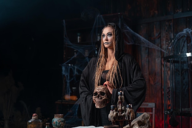 Koncepcja Halloween. Portret czarownicy z bliska z dredami trzymając w ręku ludzką czaszkę l ubrany w czarny kaptur stojący ciemny pokój widok z boku kopia przestrzeń Kaukaski nekromanta