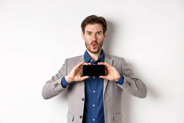 Koncepcja handlu elektronicznego i zakupów online. Podekscytowany młody człowiek w garniturze pokazuje pusty ekran smartfona i mówi wow zdziwiony, białe tło.