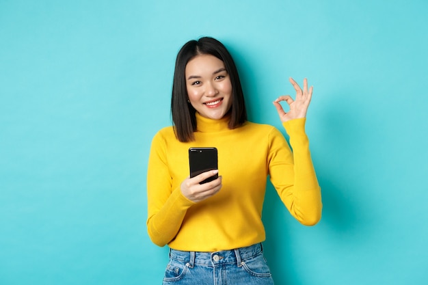 Koncepcja handlu elektronicznego i zakupów online. Portret azjatyckiej kobiety pokazującej znak OK i korzystającej z telefonu komórkowego, chwała aplikacja, stojąca na niebieskim tle.