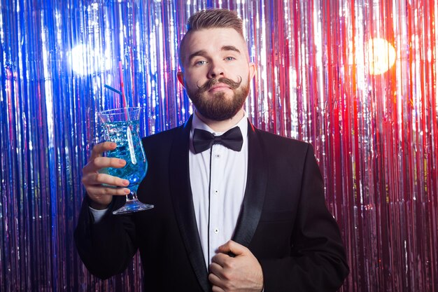 Koncepcja impreza klubowa i święta - portret elegancki przystojny mężczyzna w drogim garniturze posiada niebieski koktajl na błyszczącym tle.