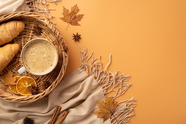 Koncepcja jesienna Zdjęcie z górnego widoku kosza z koszykiem z kubkiem piankowego kakao croissantów suszonych plasterków pomarańczowych aniz żółtych liści klonu cynamonowe patyki plaid i stożki sosnowe na izolowanym pomarańczowym tle