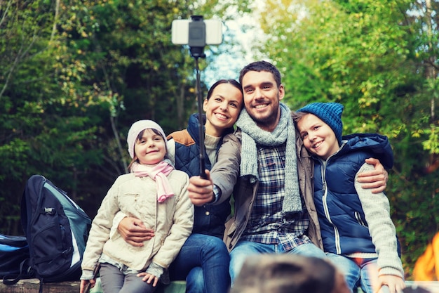 koncepcja kempingu, podróży, turystyki, wędrówek i ludzi - szczęśliwa rodzina siedzi na ławce i robi zdjęcie smartfonem na kiju selfie przy ognisku w lesie