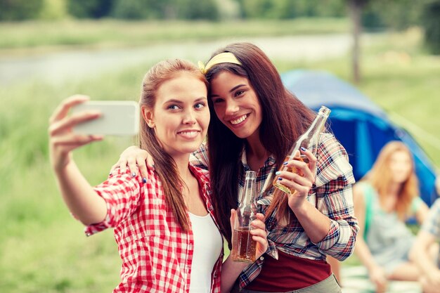 koncepcja kempingu, podróży, turystyki, wędrówek i ludzi - szczęśliwe młode kobiety ze szklanymi butelkami pijące cydr lub piwo i robiące selfie smartfonem na kempingu