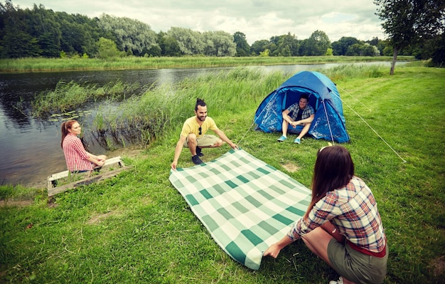 koncepcja kempingu, podróży, turystyki, wędrówek i ludzi - szczęśliwi przyjaciele z namiotem kładącym koc piknikowy na kempingu nad brzegiem rzeki