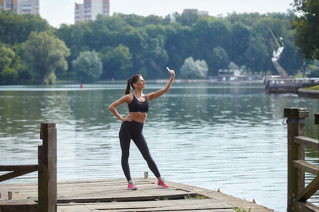 Koncepcja Kształcenia Na Odległość. Atrakcyjna Dziewczyna W Odzieży Sportowej Stoi Na Drewnianym Molo W Parku Miejskim. Lekcje Online Za Pośrednictwem Smartfona.