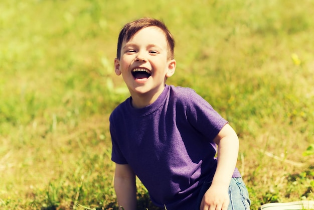 koncepcja lato, dzieciństwo, wypoczynek i ludzie - szczęśliwy mały chłopiec siedzący na trawie i śmiejący się na zewnątrz