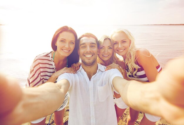 koncepcja lato, morze, turystyka, technologia i ludzie - grupa uśmiechniętych przyjaciół z aparatem na plaży, fotografująca i robiąca selfie