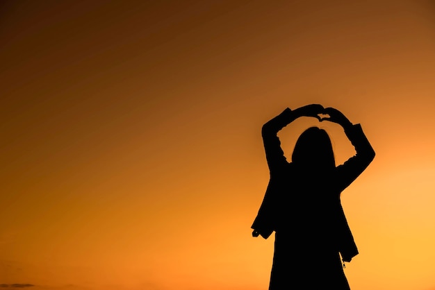 Koncepcja lub pojęciowy kształt serca lub symbol wykonany z kobiety ręka sylwetka na niebie na tle zachodu słońca dla miłości