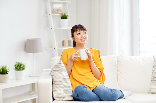 koncepcja ludzi, napojów i wypoczynku - szczęśliwa azjatycka kobieta siedząca na kanapie i pijąca herbatę z filiżanki lub kubka w domu